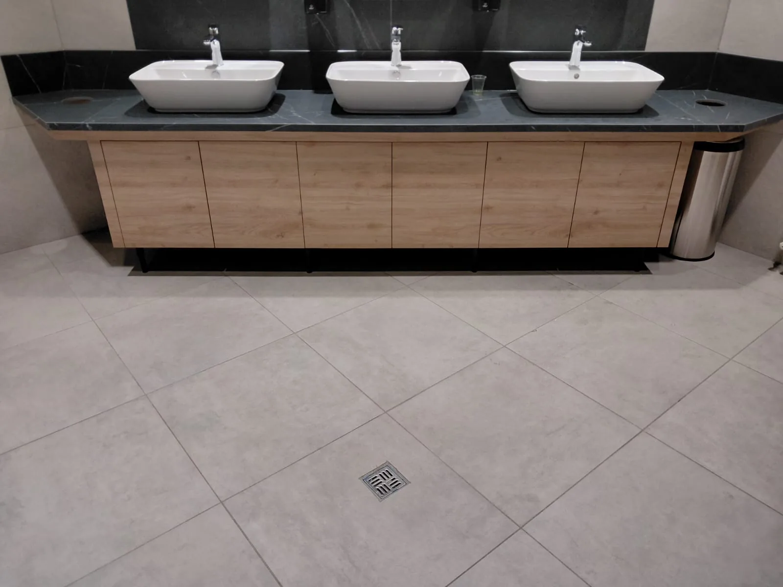Koyu damarlı tezgah üzerinde üç beyaz lavabo ve siyah ayaklı, ahşap görünümlü altı kapaklı dolap